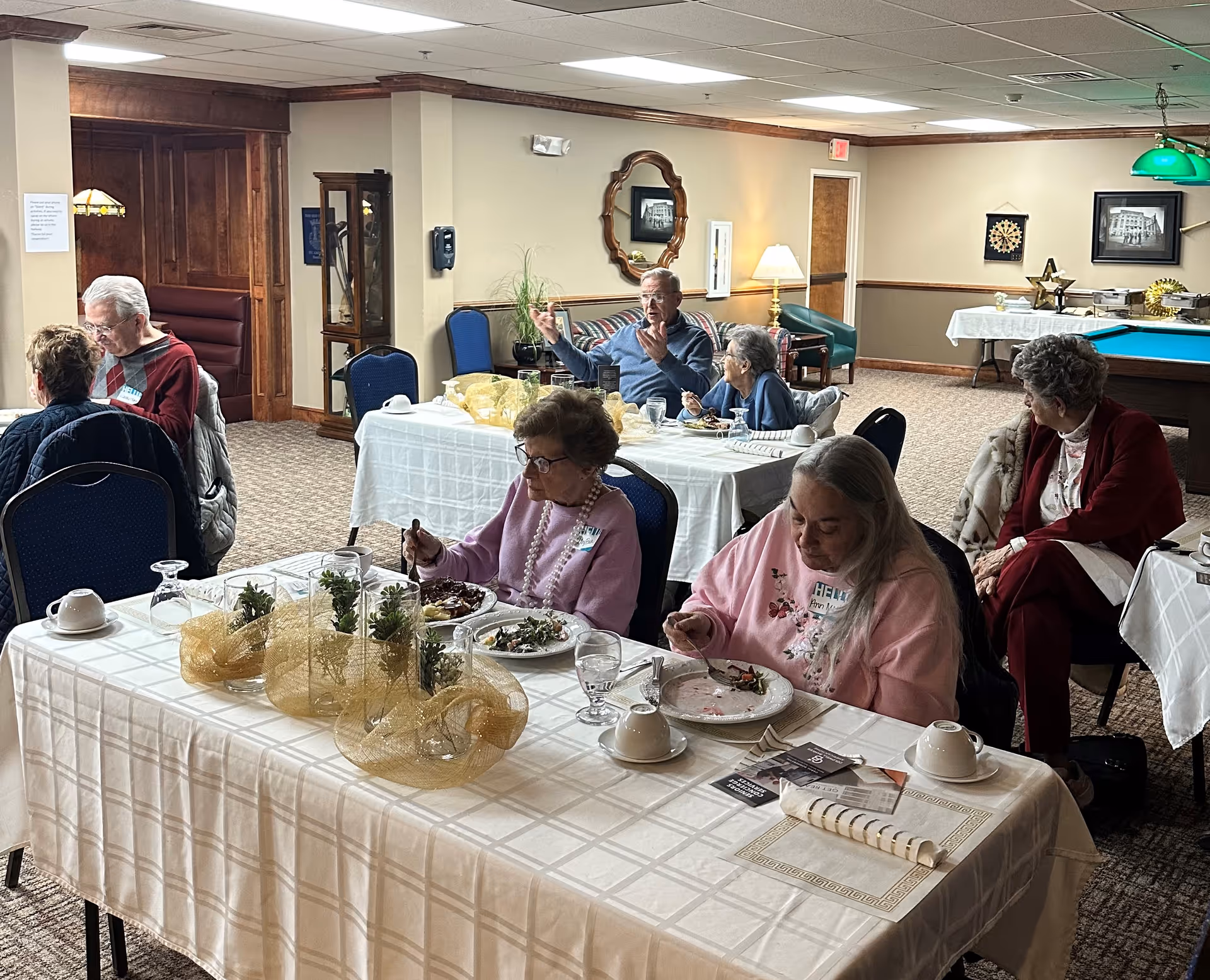 Several elderly residents sit at decorated tables eating and conversing in a dining/recreation room with a pool table in the background.