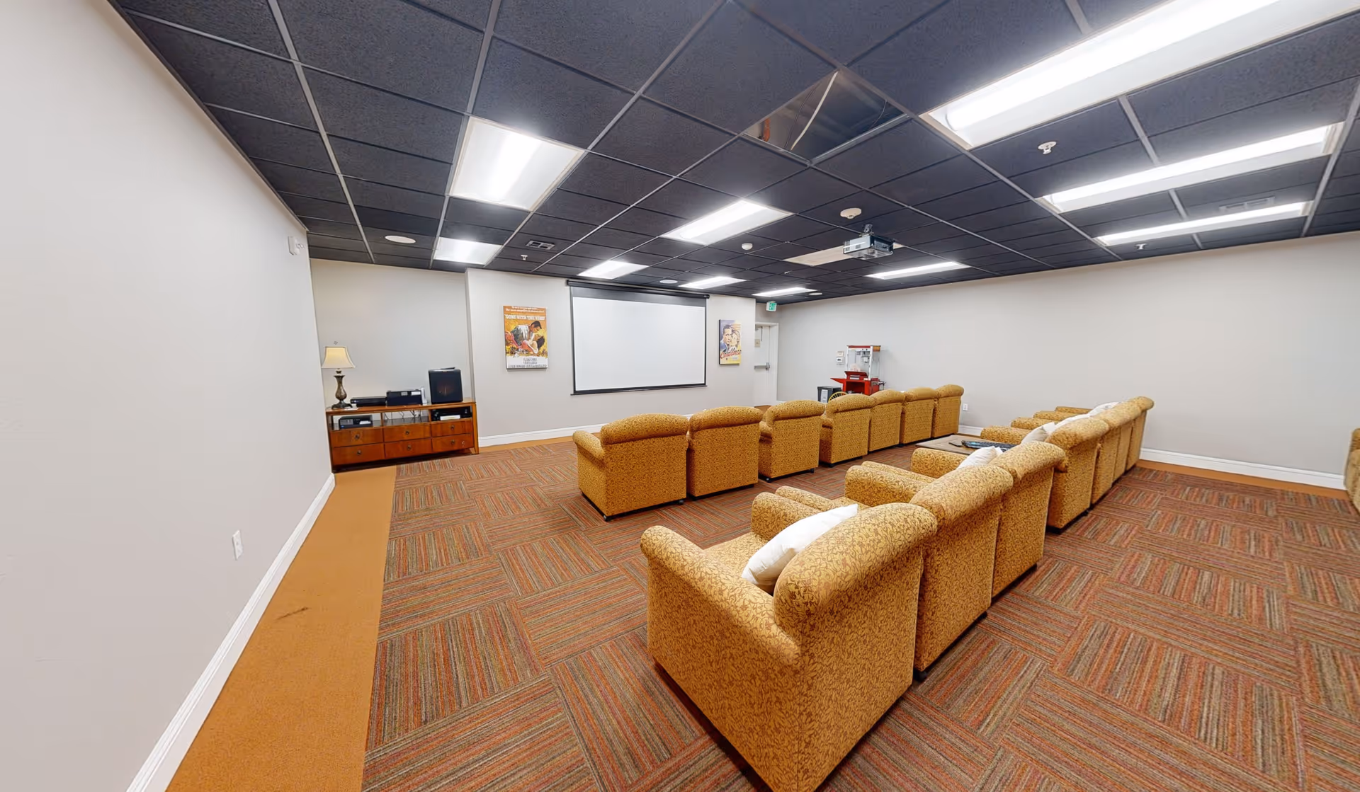 A small theater room with two rows of cushioned armchairs facing a large white projection screen on the wall. The room has a patterned carpet, a black ceiling with recessed lighting, and a wooden cabinet with a lamp and audio equipment on the left side. There is also a popcorn machine in the back corner.