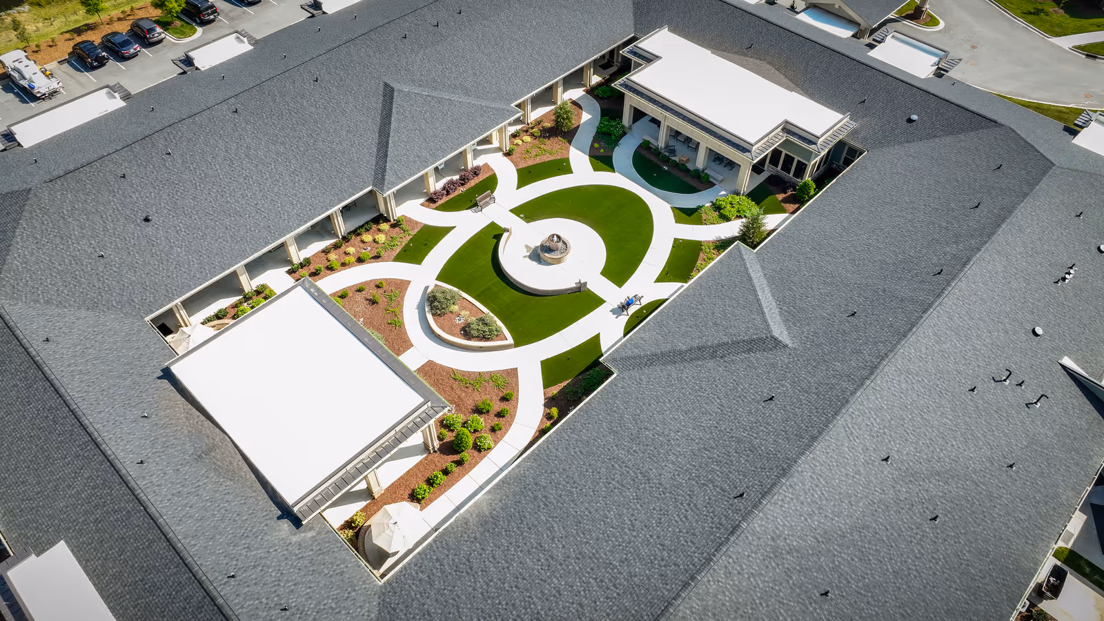 Aerial view of a senior living facility courtyard with a circular walking path, green lawn areas, landscaped garden beds, and a central fountain. The courtyard is surrounded by a building with a gray roof and covered walkways.