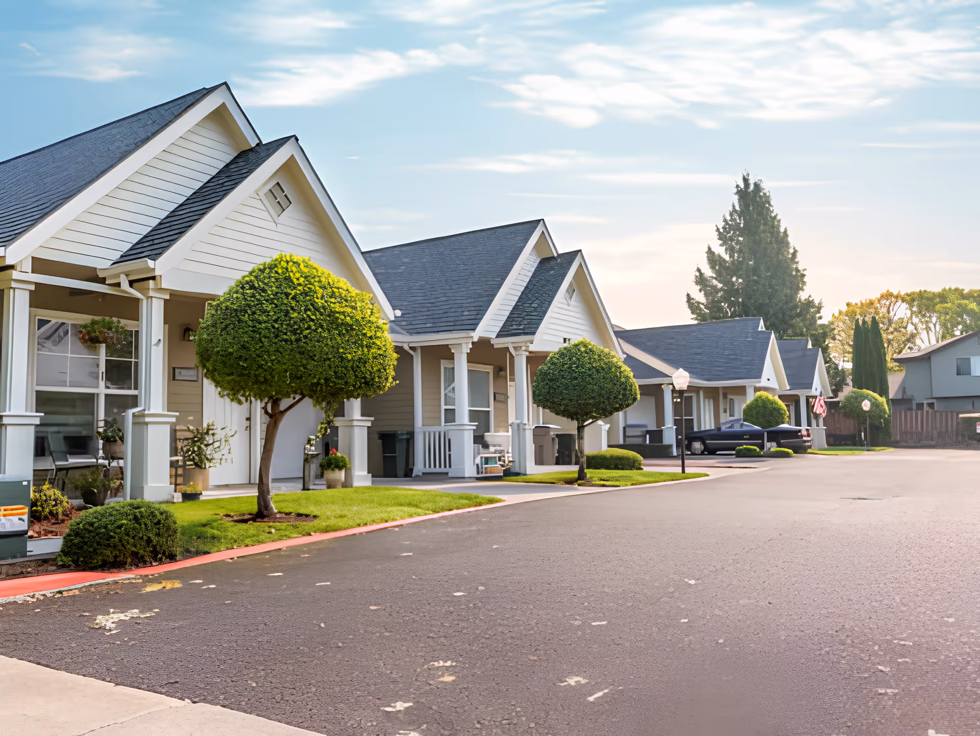 Row of single-story residential buildings with white siding and dark roofs, each with a small porch and neatly trimmed round bushes in front, along a paved driveway under a partly cloudy sky.