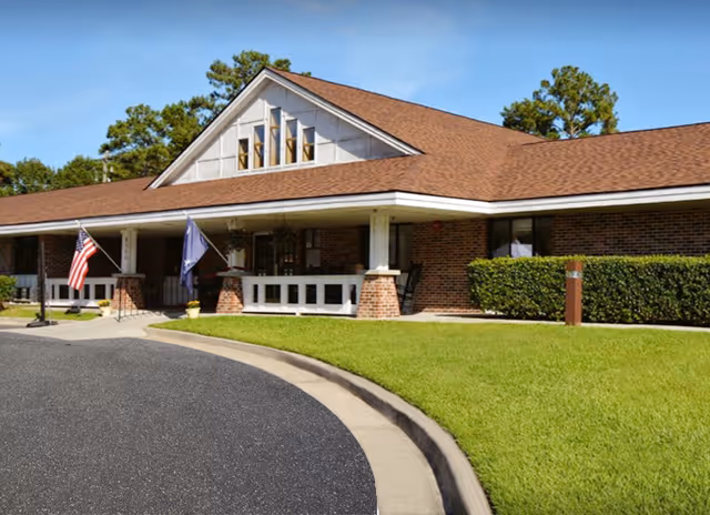 Exterior view of a single-story brick building with a brown shingled roof, a covered porch with white railings, two flags displayed near the entrance, and well-maintained green lawn and shrubs under a clear blue sky.
