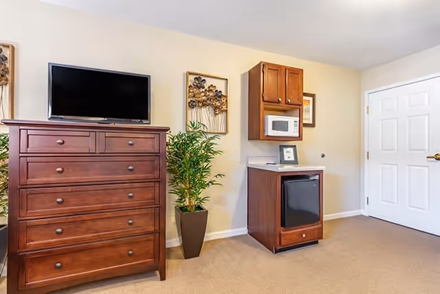 Interior view of a room featuring a wooden dresser with a flat-screen TV on top, a tall potted plant beside it, and a small kitchenette area with a microwave, mini refrigerator, and wooden cabinets mounted on the wall. The room has beige walls and carpeted flooring, with a white door on the right side.