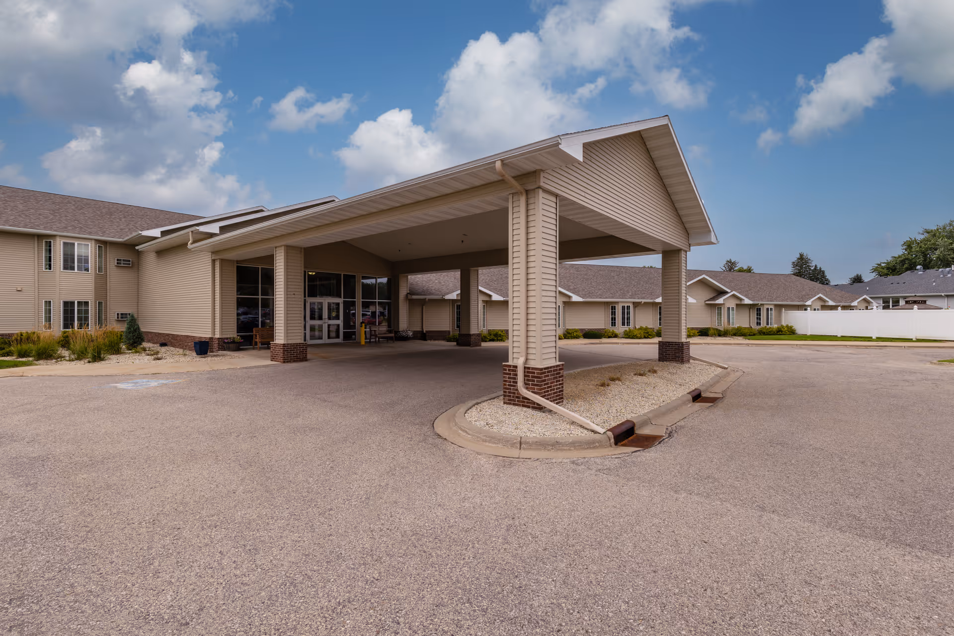 Exterior view of Prairie Meadows Senior Living facility showing the main entrance with a covered drop-off area supported by columns, surrounded by a paved driveway and landscaped with small bushes and rocks under a partly cloudy sky.