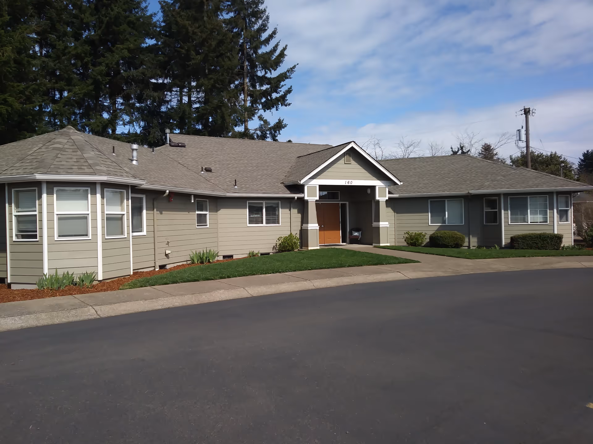 Exterior view of a single-story building with gray siding and a gable roof, surrounded by green grass and bushes, with tall trees in the background under a partly cloudy sky.