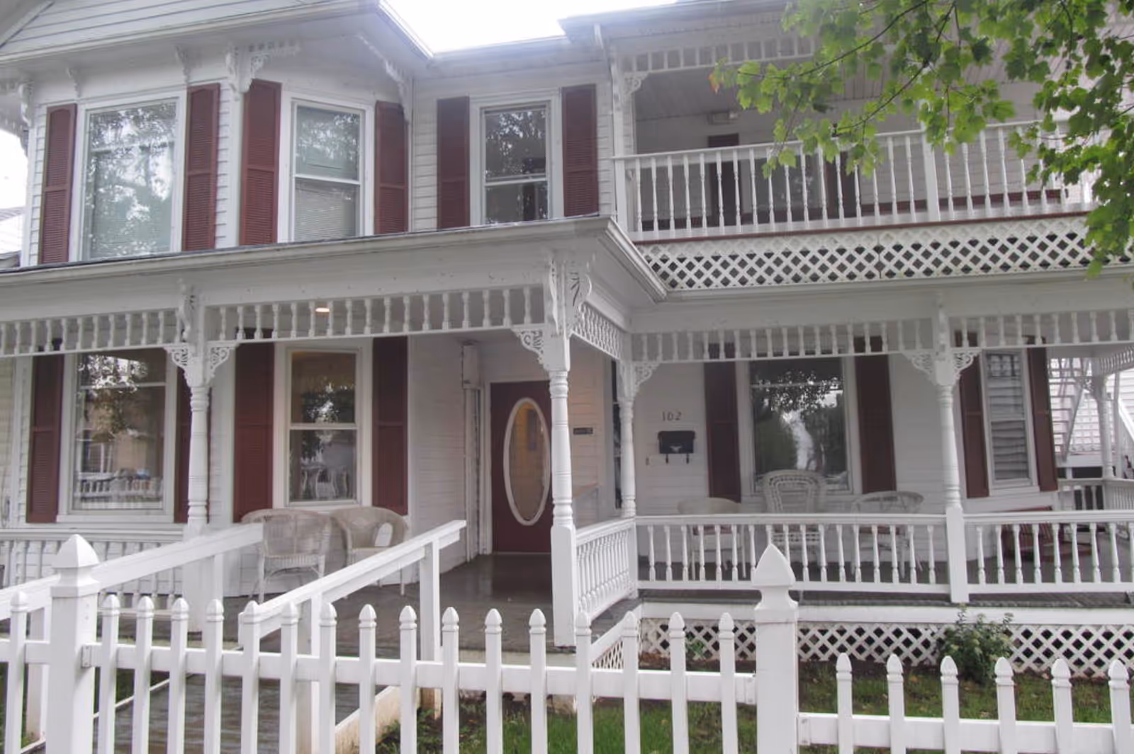 Front view of a two-story white Victorian-style house with a wraparound porch, white picket fence, and red shutters.