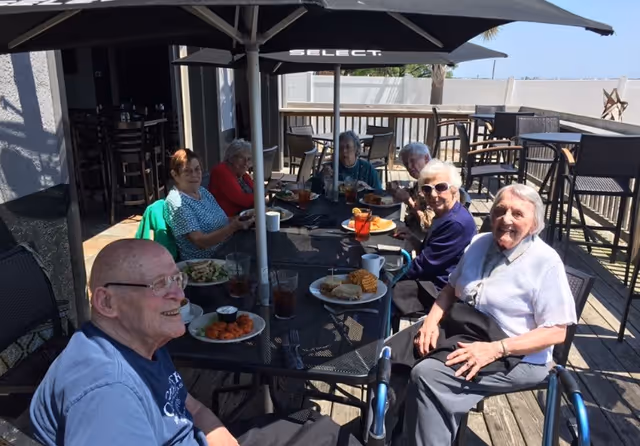 A group of elderly people sitting around a black metal outdoor table under a large umbrella, enjoying a meal together on a wooden deck. Plates of food and drinks are on the table, and the setting appears sunny and relaxed.