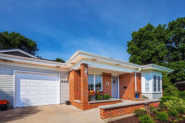 Exterior view of a single-story brick and siding residential building with a white garage door, a small porch with potted plants, and a well-maintained garden under a clear blue sky.