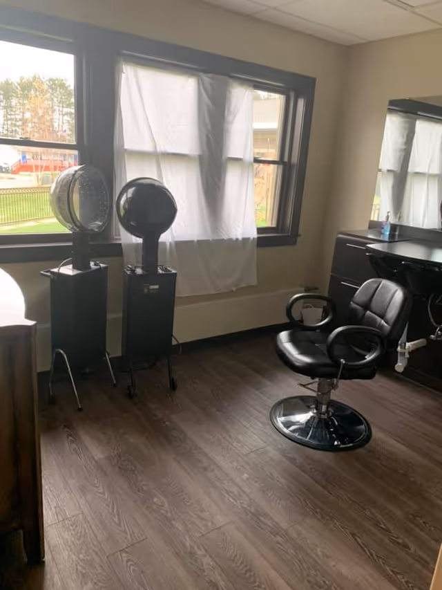 Interior salon room with a styling chair, hooded hair dryers, a sink station and a window covered by a sheet.