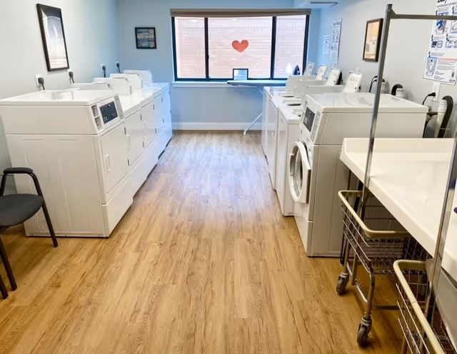 A clean communal laundry room with rows of washers and dryers, folding tables and laundry carts, and a window with a heart decoration.