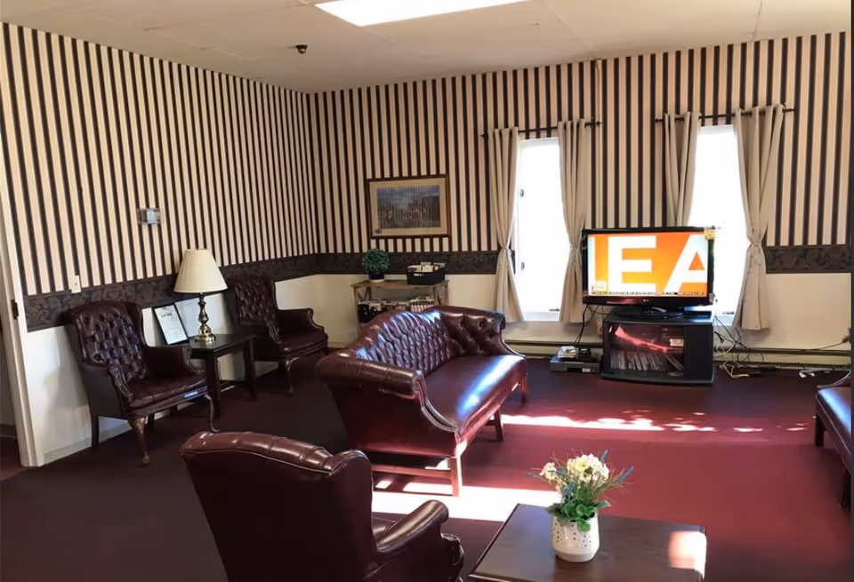 A living room with burgundy leather chairs and a matching loveseat arranged around a coffee table with a small flower arrangement. The walls have vertical striped wallpaper with a dark patterned border. Two windows with beige curtains let in natural light. A flat-screen TV is placed on a stand between the windows.