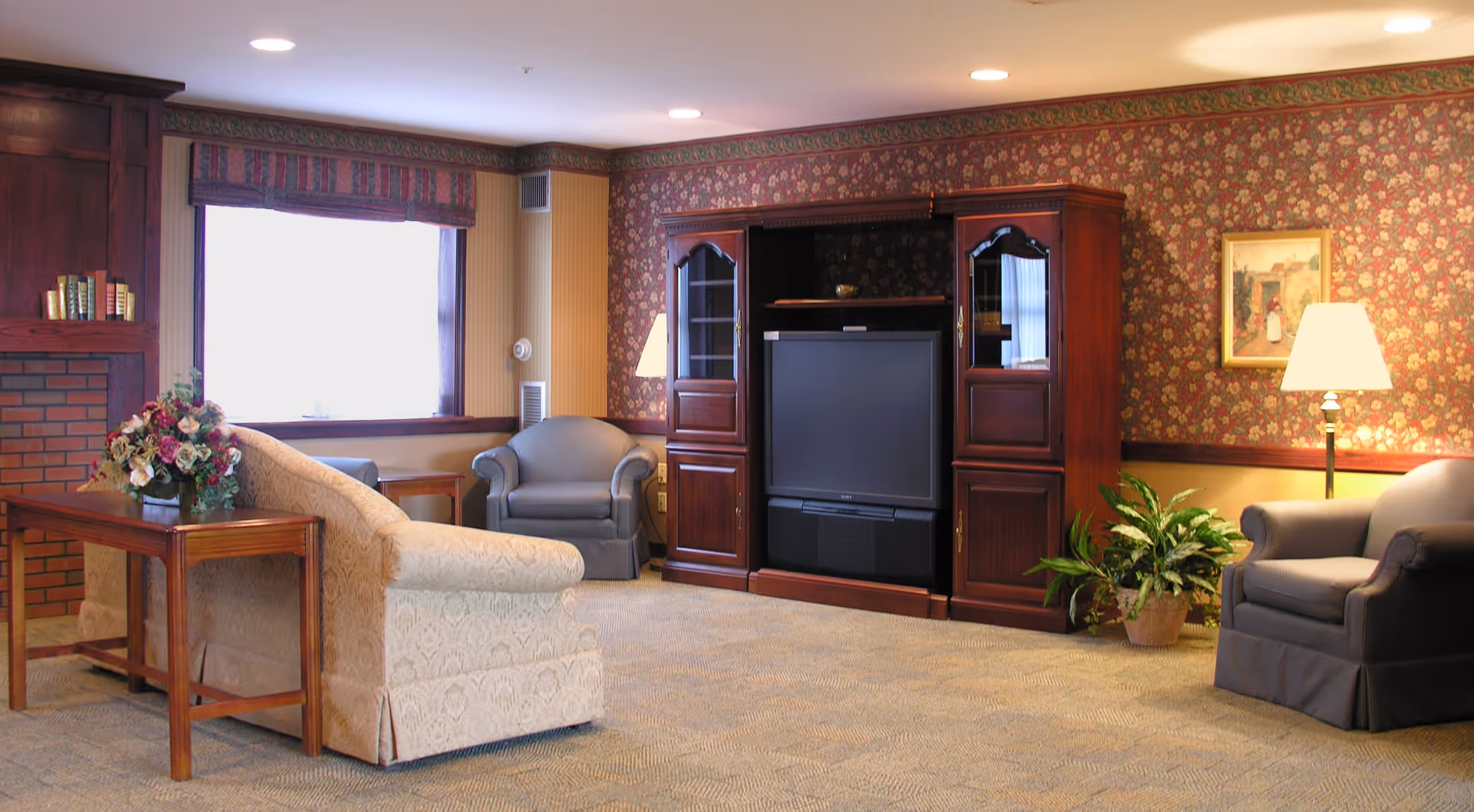 A cozy living room with floral wallpaper and carpeted floor. The room features a beige patterned sofa, two gray armchairs, a wooden entertainment center with a television, a wooden side table with a floral arrangement, a floor lamp, a potted plant, and a framed painting on the wall.