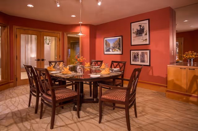 A warmly lit dining room with a round wooden table set for six people. The table has place settings with plates, glasses, and folded yellow napkins. The chairs have patterned cushions. The walls are painted a deep red and decorated with framed pictures. There are double wooden doors with frosted glass panels and a sideboard with a vase of flowers.
