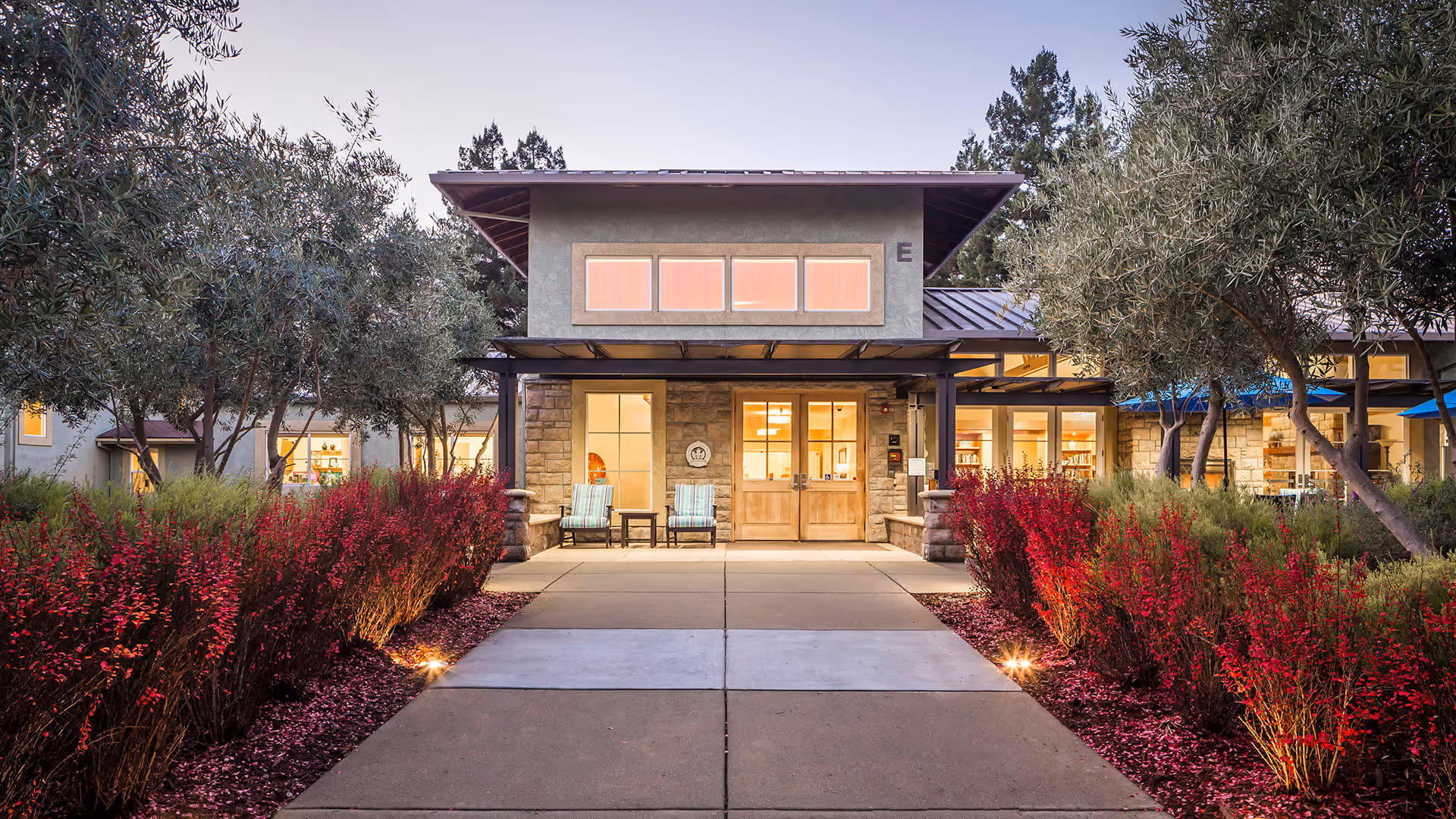 Entrance to a senior living facility building with a paved walkway flanked by red flowering bushes and trees. The building has large windows and double wooden doors, with two chairs and a small table on the porch area. The scene is captured during dusk with warm interior lights visible through the windows.