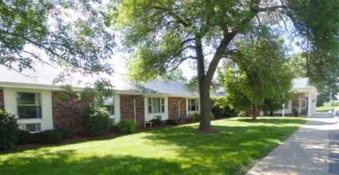 Single-story brick senior living facility with green lawn, trees, and a sidewalk.