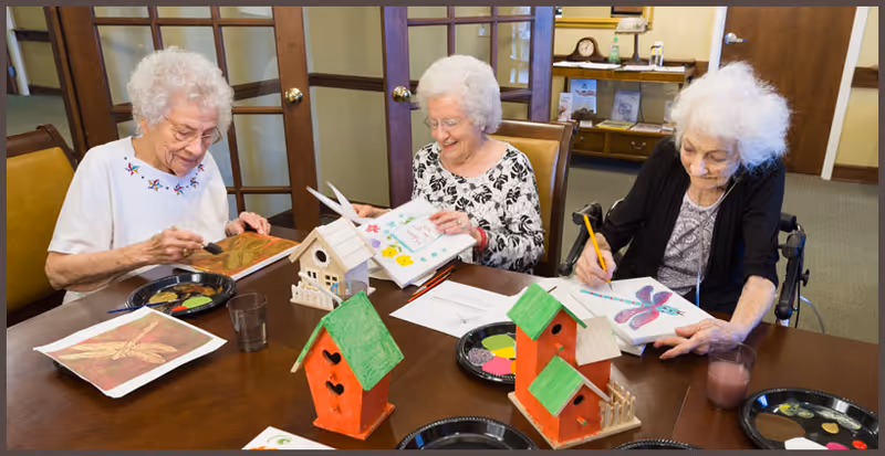 Three elderly women sitting around a table engaged in arts and crafts activities, painting and drawing on paper and wooden birdhouses in a well-lit room with wooden furniture and glass-paneled doors.