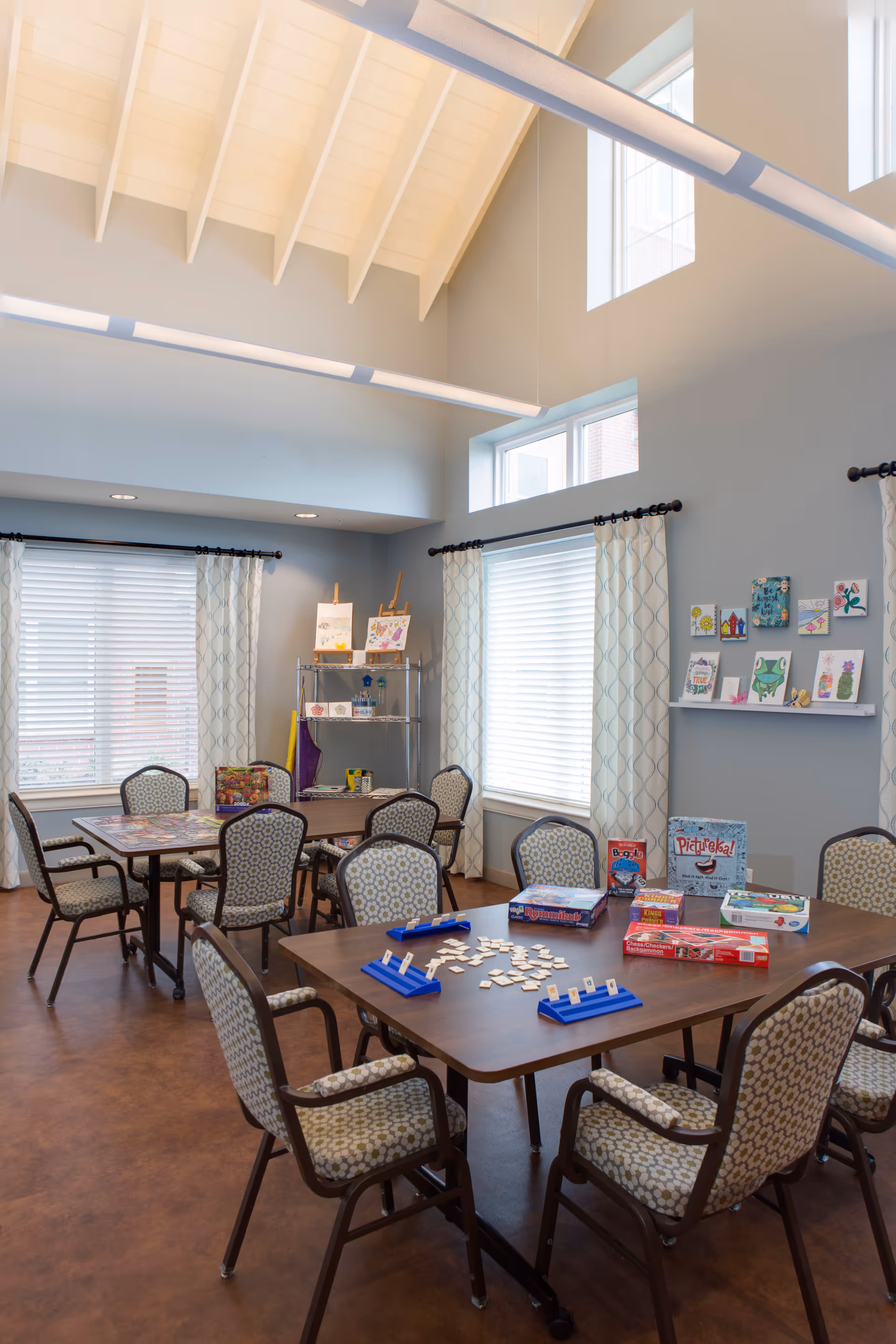 A bright activity room with high vaulted ceilings and large windows covered with patterned curtains. Several tables and cushioned chairs are arranged around the room. On the tables are various board games and game pieces. The walls have shelves displaying colorful artwork and crafts. The floor is a warm brown color, and the room has a welcoming, organized atmosphere.