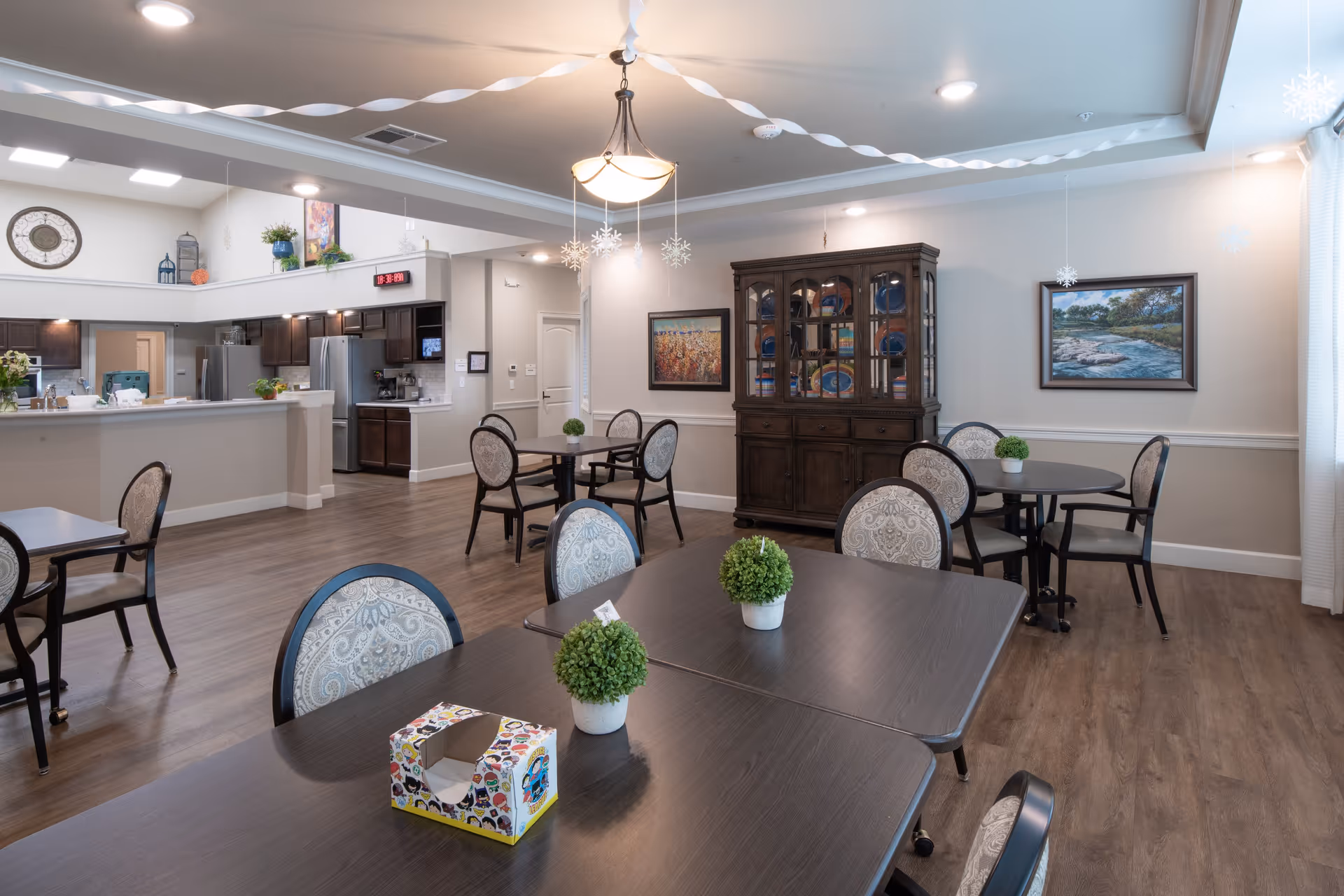 A bright and spacious dining area in an assisted living facility featuring several dark wood tables with patterned cushioned chairs. Small green potted plants decorate the tables. The room has wood flooring, light-colored walls, and ceiling lights. A dark wood china cabinet with decorative plates is against one wall, flanked by framed landscape paintings. The kitchen area with stainless steel appliances and dark cabinetry is visible in the background. White paper streamers and hanging snowflake decorations adorn the ceiling.