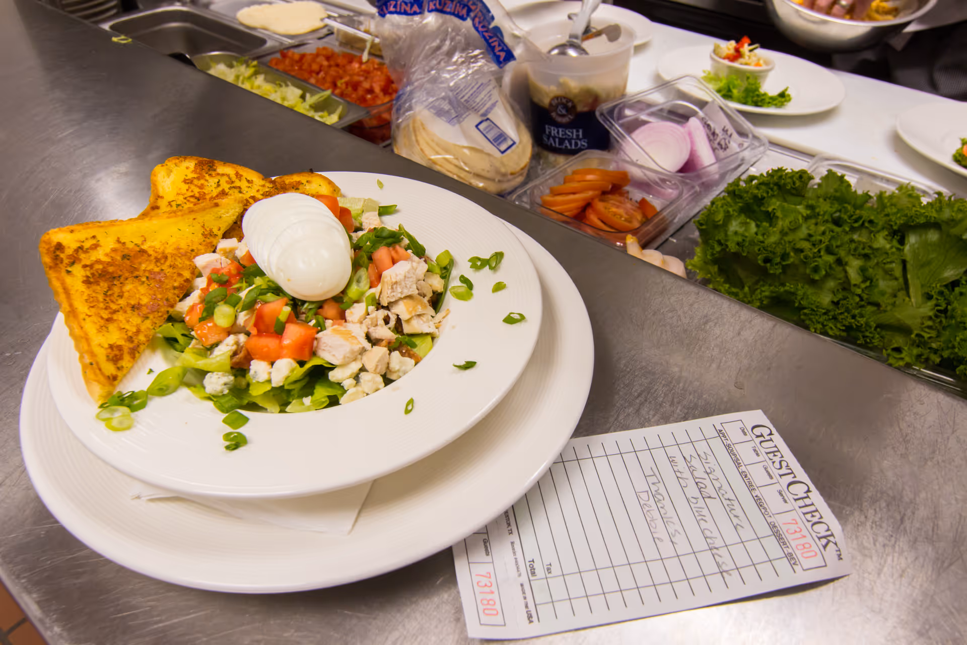 A plated salad with chopped chicken, diced tomatoes, green onions, blue cheese crumbles, and a sliced hard-boiled egg on top, served with two pieces of toasted bread. The plate is on a kitchen counter with various salad ingredients like lettuce, sliced tomatoes, onions, and tortillas in the background. A guest check slip is also visible on the counter.