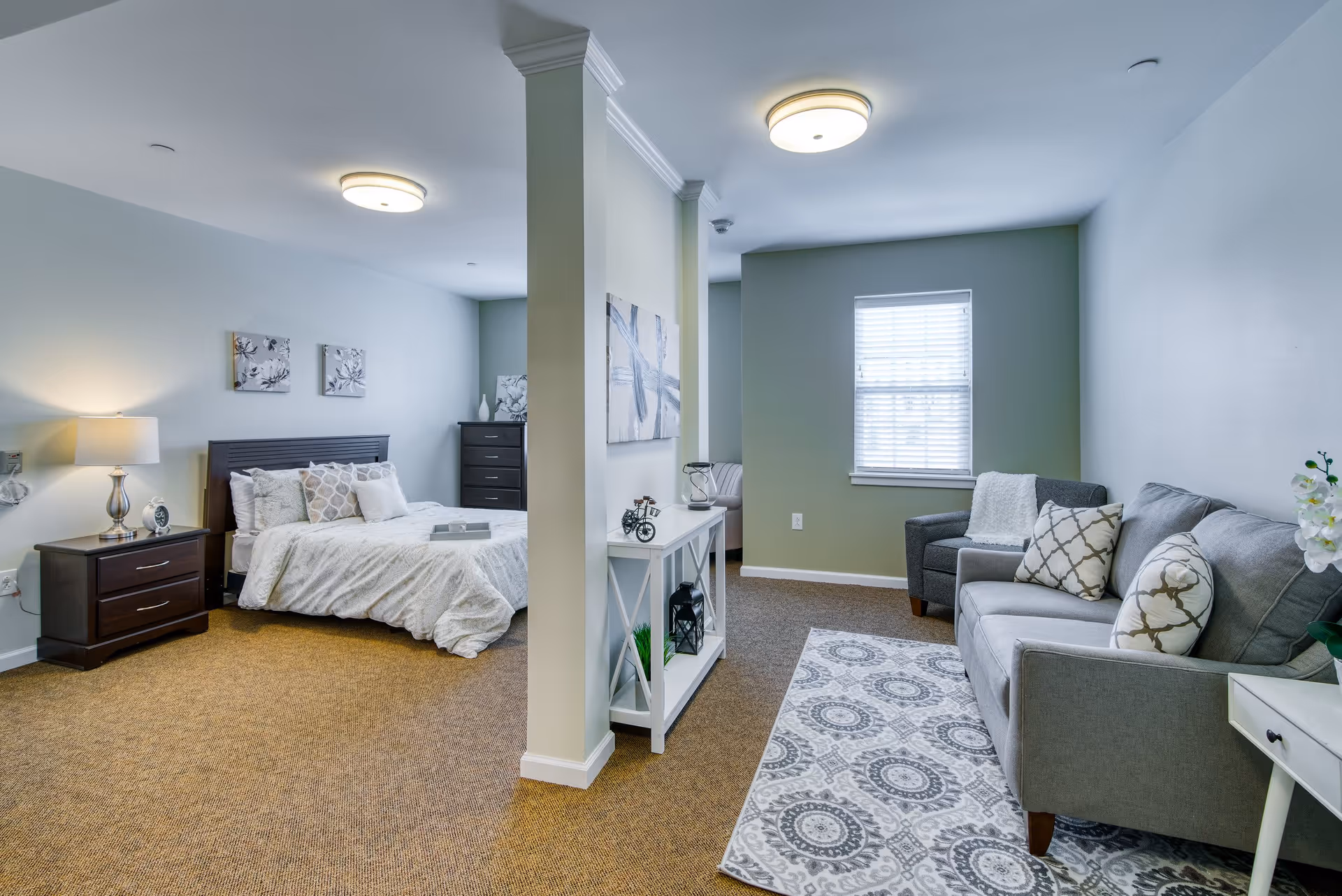 Studio-style senior living interior with a bed and nightstand on the left and a seating area with sofa and armchair on the right separated by a partial wall.