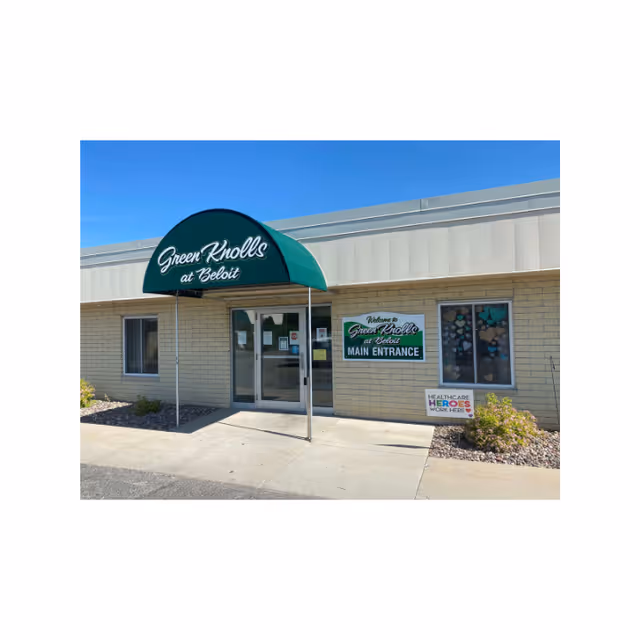 The main entrance of Green Knolls at Beloit with a green awning over glass doors and a 'Main Entrance' sign on a beige brick facade under a clear blue sky.