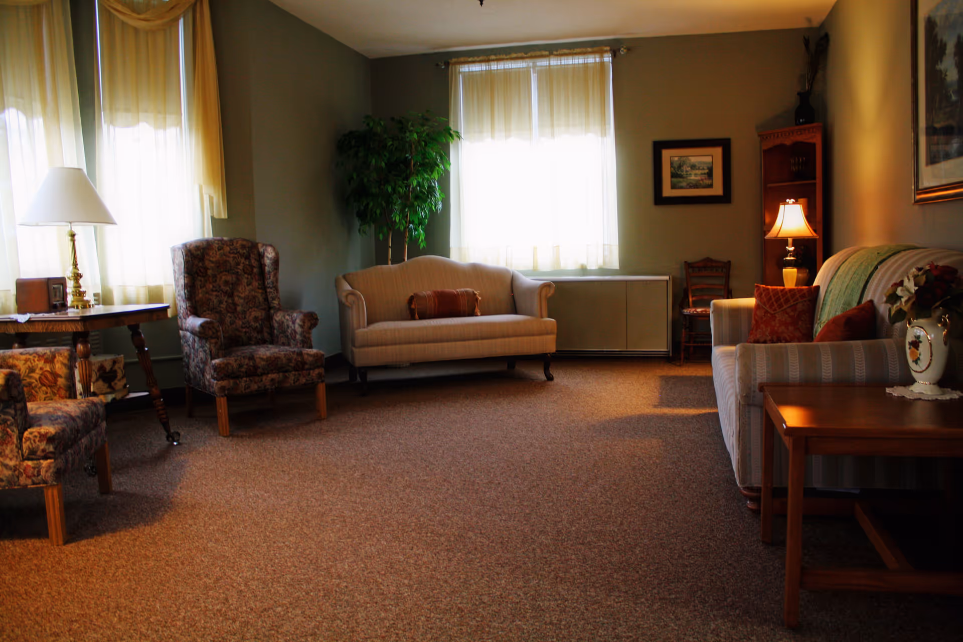 A cozy living room with beige carpet, two patterned armchairs, a beige loveseat with a decorative pillow, a striped sofa with red and green pillows, a wooden side table with a lamp and a vase of flowers, a wooden corner shelf with a lamp, framed artwork on the walls, and two windows with sheer curtains letting in natural light.