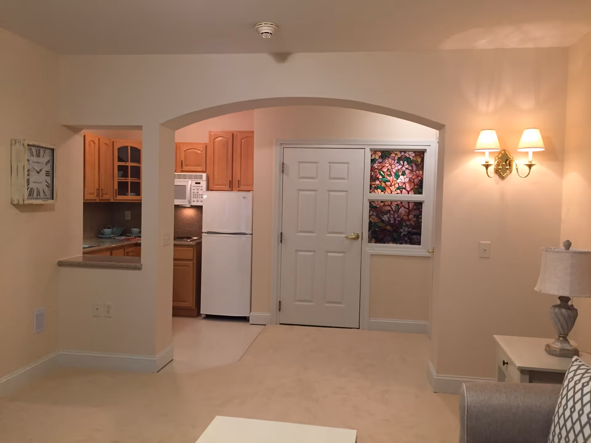 Interior view of a senior living facility showing a small kitchen area with wooden cabinets, a white refrigerator, and a microwave. The kitchen is visible through an arched opening from a carpeted living area with a side table, lamp, and wall-mounted double light fixture. A white door with decorative stained glass panels is also visible.