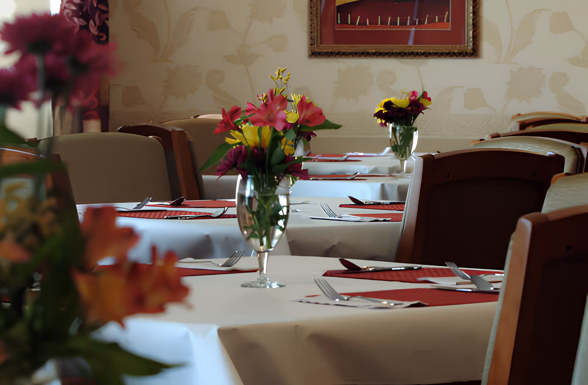 A dining room with tables covered in white tablecloths and red placemats, each set with silverware and a glass vase holding colorful flowers. Wooden chairs surround the tables, and a framed picture hangs on the wall with floral wallpaper in the background.