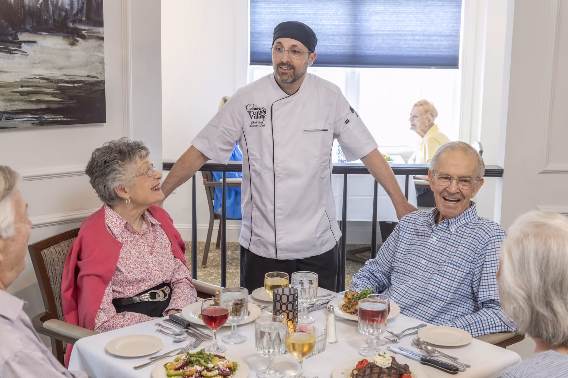 A chef wearing a white uniform and black hat stands and talks with four elderly people seated around a dining table set with plates of food and glasses of beverages in a bright dining room.