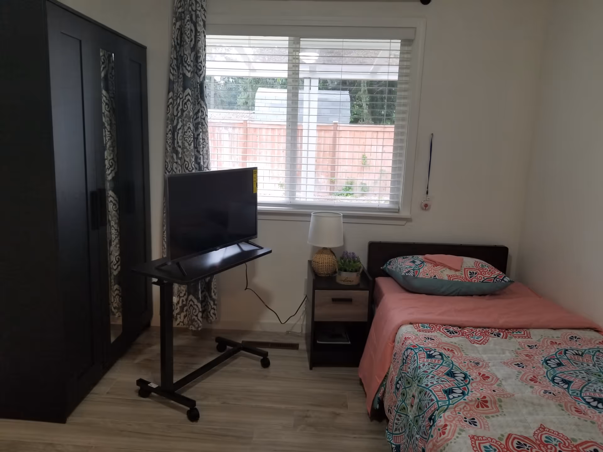 Small furnished bedroom with a single bed, nightstand and lamp, TV on a rolling stand, and a wardrobe by a window.