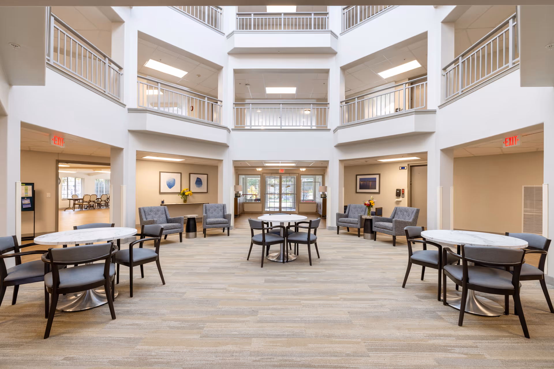 A spacious, well-lit common area in Holiday Kittery Estates featuring round tables with chairs and several armchairs arranged along the walls. The area has a high ceiling with balconies on the upper floors and large windows letting in natural light. There are exit signs visible above doorways on both sides.