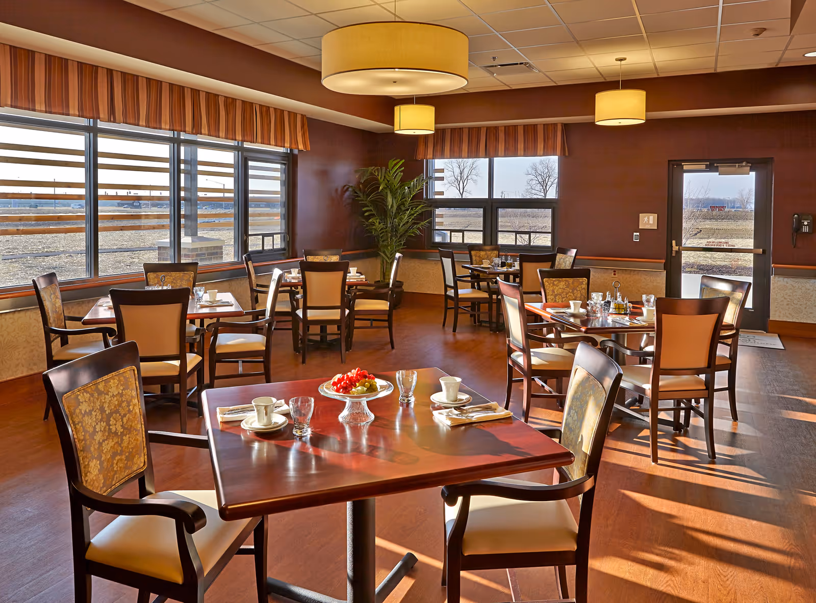 Sunlit dining room with several set tables and chairs, large windows, hanging lights, and a bowl of fruit on the nearest table.