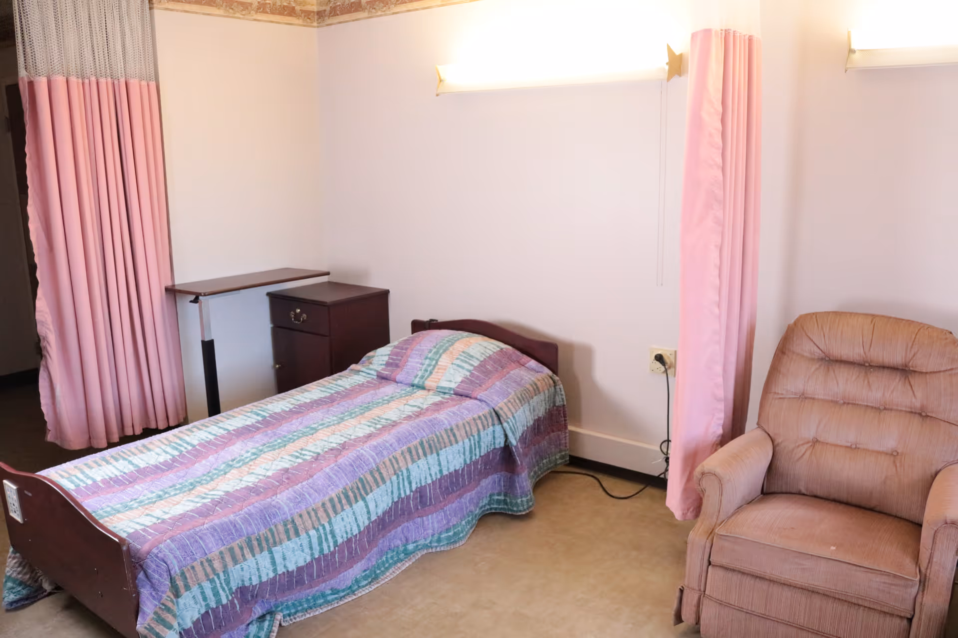 A simple room with a single bed covered with a striped quilt in shades of purple, blue, and green. Next to the bed is a small dark wooden nightstand and an adjustable overbed table. Pink privacy curtains hang on either side of the bed. To the right of the bed is a cushioned beige recliner chair. The walls are light-colored and the floor is a neutral tone.
