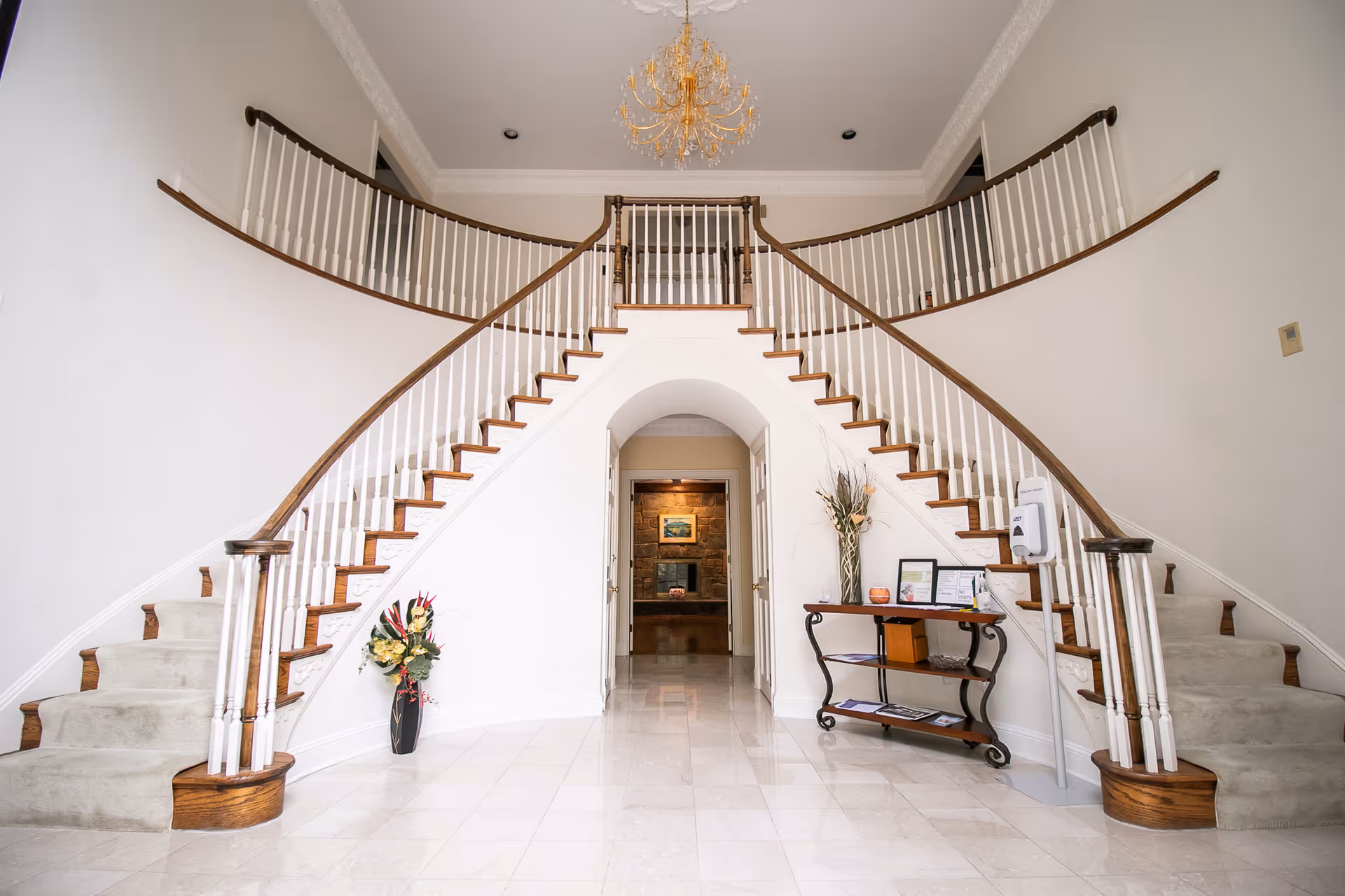 A grand interior entrance featuring a double staircase with wooden handrails and white balusters curving upwards on both sides. The floor is tiled in light-colored marble, and a chandelier hangs from the ceiling. There is a small table with decorative items and framed certificates on the right side, and a vase with flowers on the left side near the base of the stairs. An arched doorway leads to another room with a stone wall and a painting visible in the background.