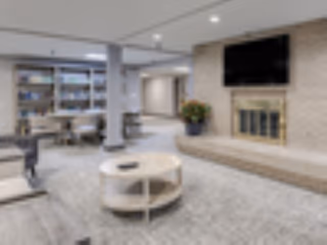 Carpeted communal living room with seating, a round coffee table, a fireplace with a TV above, and bookshelves in the background.