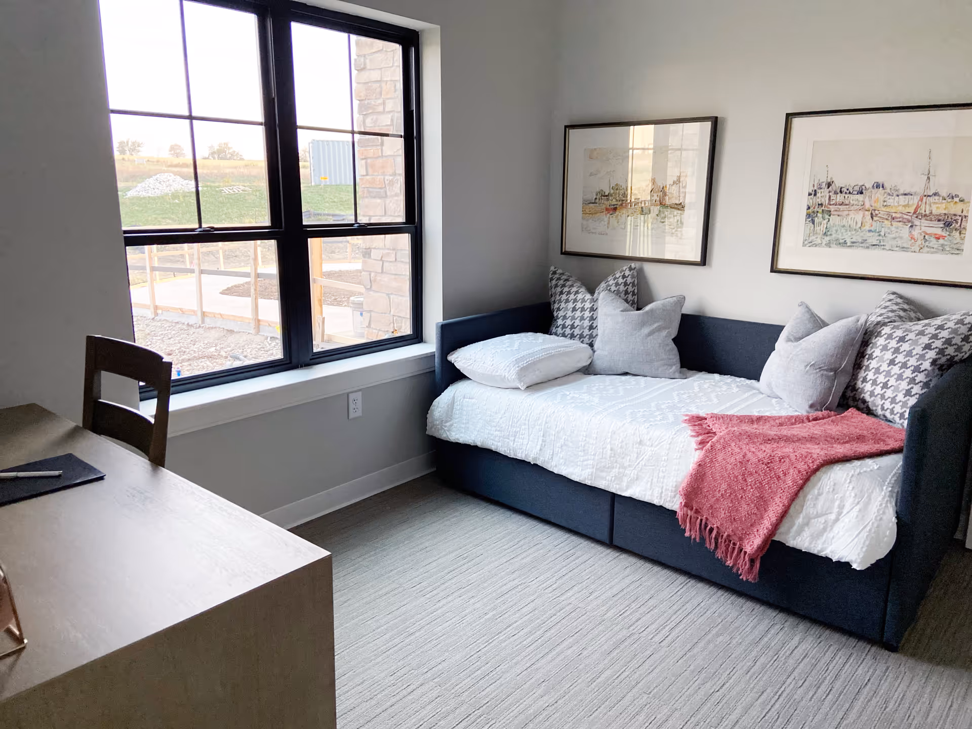 A cozy bedroom with a daybed covered in white bedding and multiple pillows, including patterned and solid gray ones. A red throw blanket is draped over the bed. Two framed watercolor paintings hang on the wall above the bed. A wooden desk and chair are positioned near a large window that lets in natural light, showing an outdoor view with grass and a stone wall.