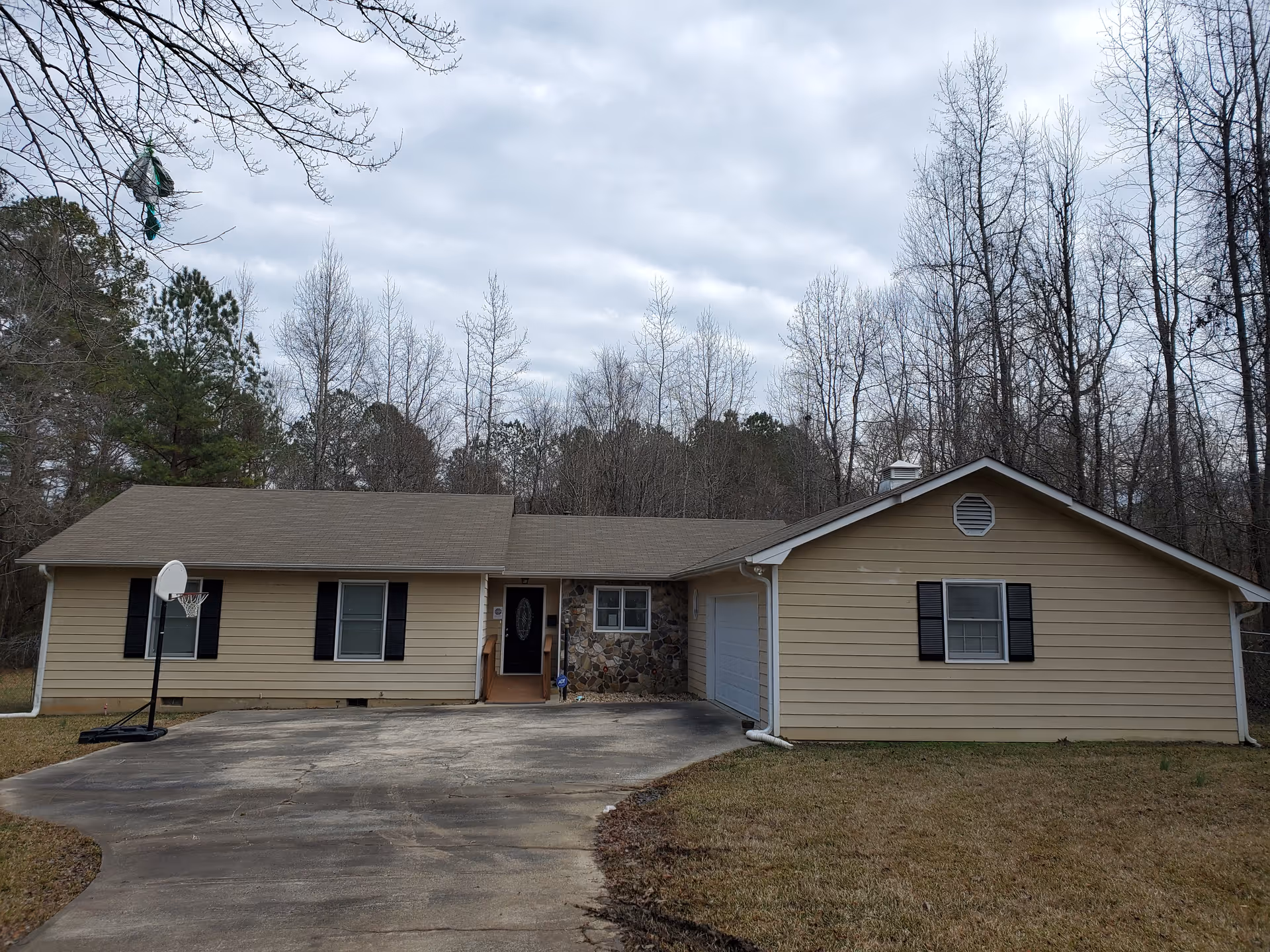 Single-story beige ranch-style house with an attached garage, driveway and a portable basketball hoop, set against leafless trees under a cloudy sky.