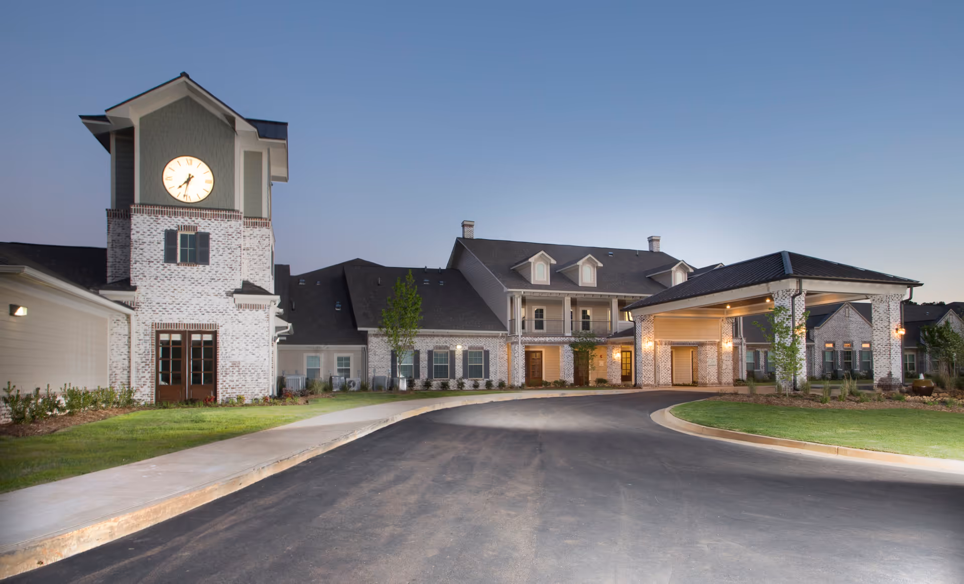 Exterior view of The Claiborne at McComb senior living facility at dusk, featuring a large brick building with a clock tower, covered entrance, and well-maintained landscaping.