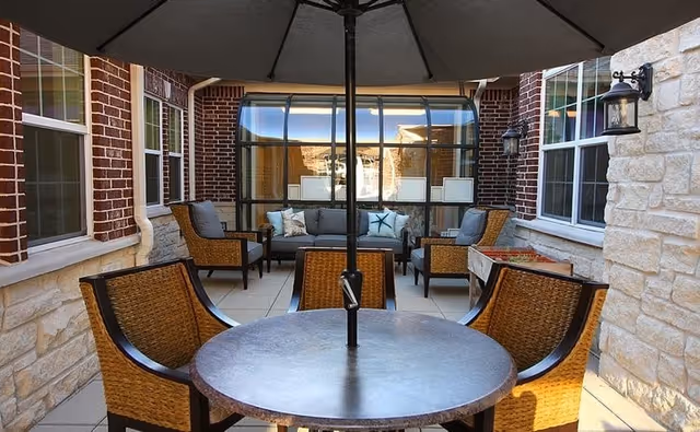 Outdoor patio area with a round table and four wicker chairs under a large umbrella. The patio is surrounded by brick and stone walls with windows and outdoor wall lanterns. In the background, there is a seating area with a sofa and two armchairs with cushions.