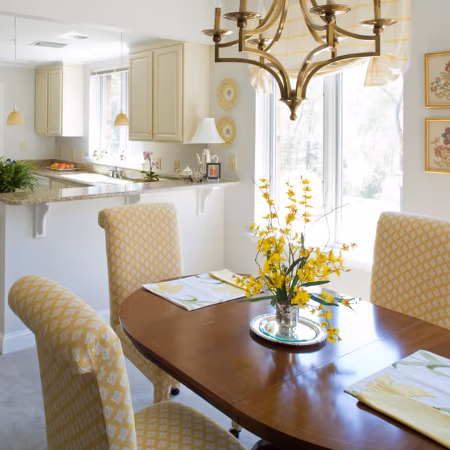 A bright dining area with a wooden table set with yellow floral placemats and a silver vase holding yellow flowers. Four upholstered chairs with a yellow and white diamond pattern surround the table. A large window with a yellow checkered valance lets in natural light. In the background, a kitchen with cream-colored cabinets, granite countertops, and pendant lights is visible.