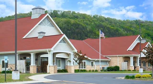 Single-story senior living facility entrance with red gabled roofs, white columns, and an American flag in front of a wooded hillside.