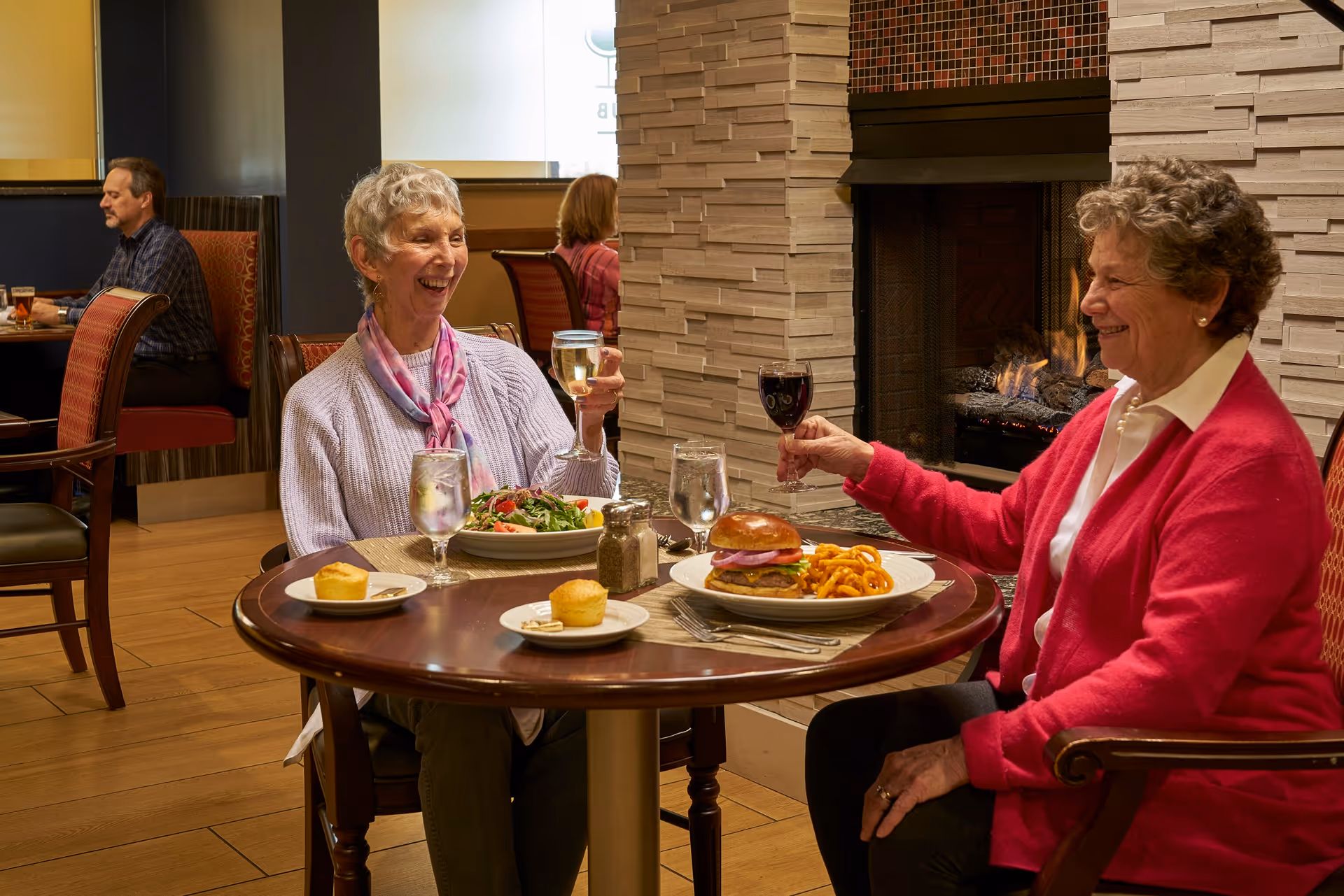 Two women smiling and clinking wine glasses across a table with plates of food in a cozy dining area near a fireplace.