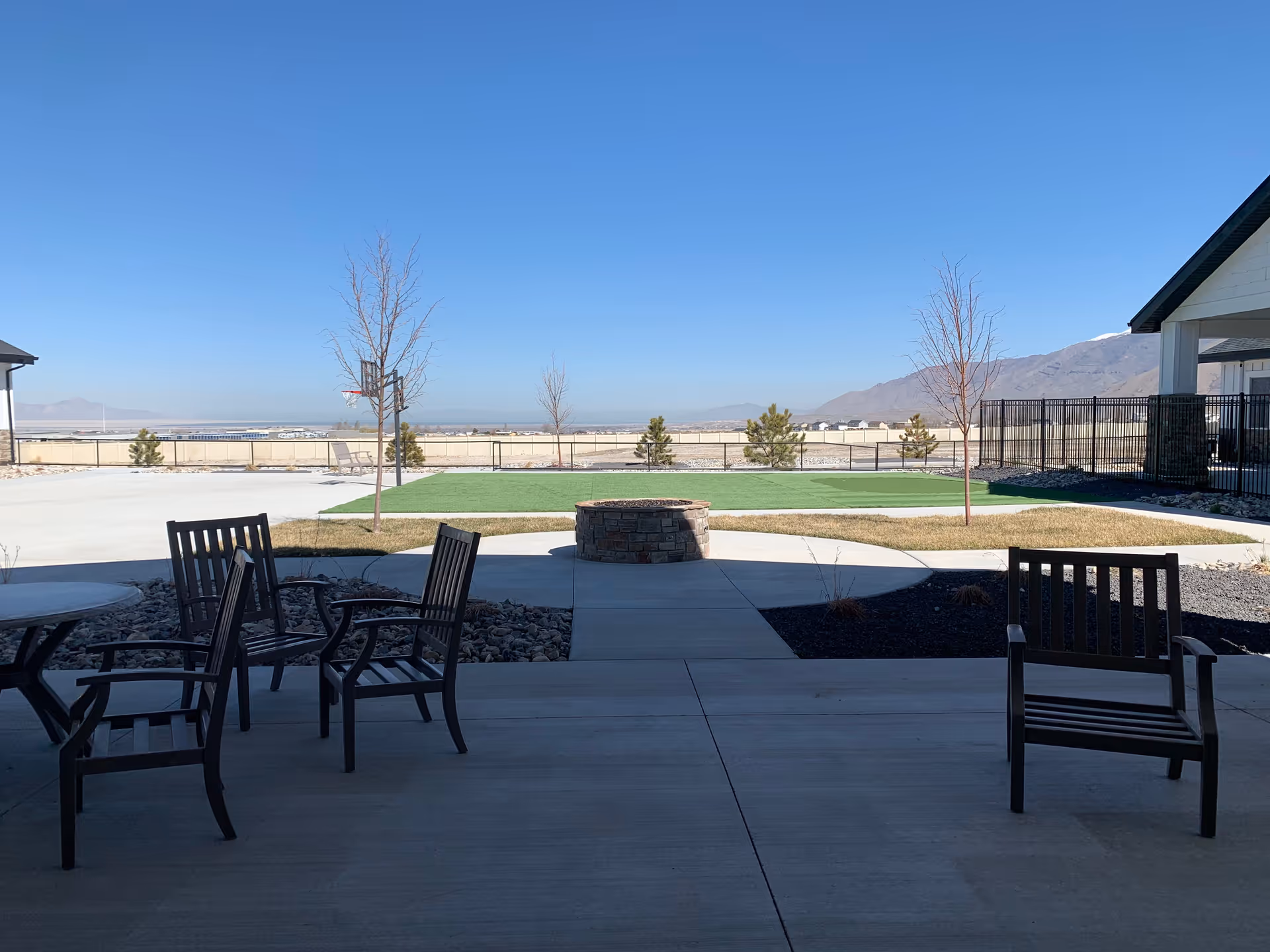 Outdoor patio area with several wooden chairs and a round table on a concrete surface, a circular stone fire pit in the center, small trees, a fenced grassy area, and mountains in the background under a clear blue sky.