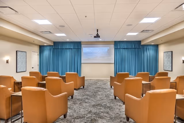 A small theater-style activity room with rows of leather armchairs facing a projection screen framed by blue curtains.