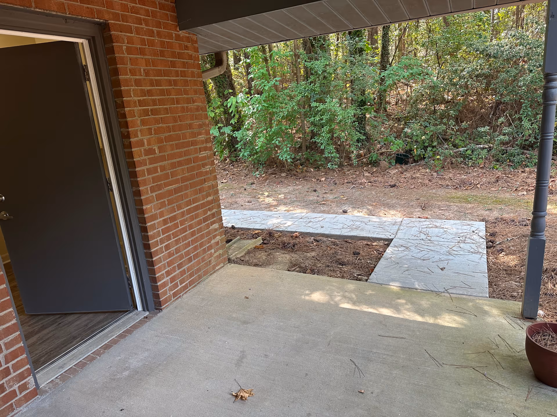 View of a covered outdoor patio area with a concrete floor and a brick wall on the left side. An open door leads inside to a room with wood flooring. Beyond the patio, there is a concrete walkway and a wooded area with green trees and bushes.