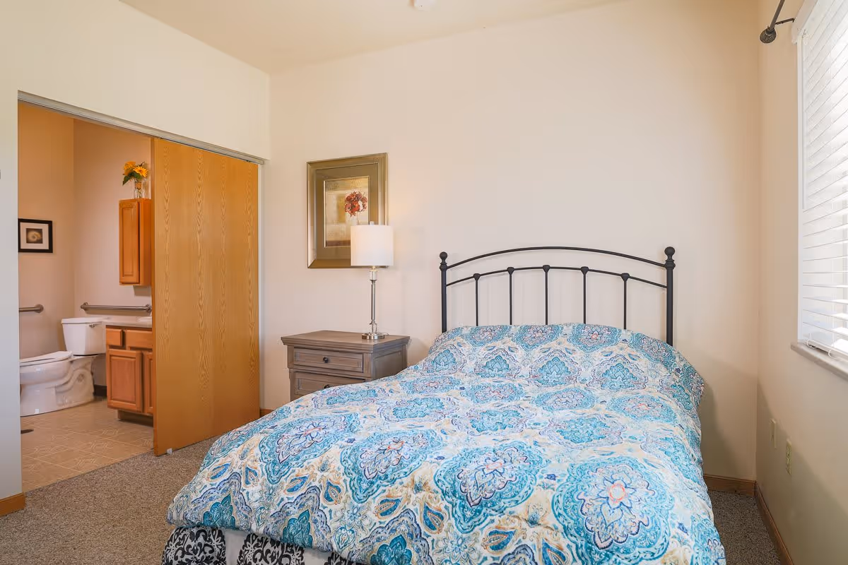 A bedroom with a bed covered in a blue and white patterned comforter, a nightstand with a lamp, a framed picture on the wall, and an open sliding door revealing a bathroom with a toilet and wooden cabinets.