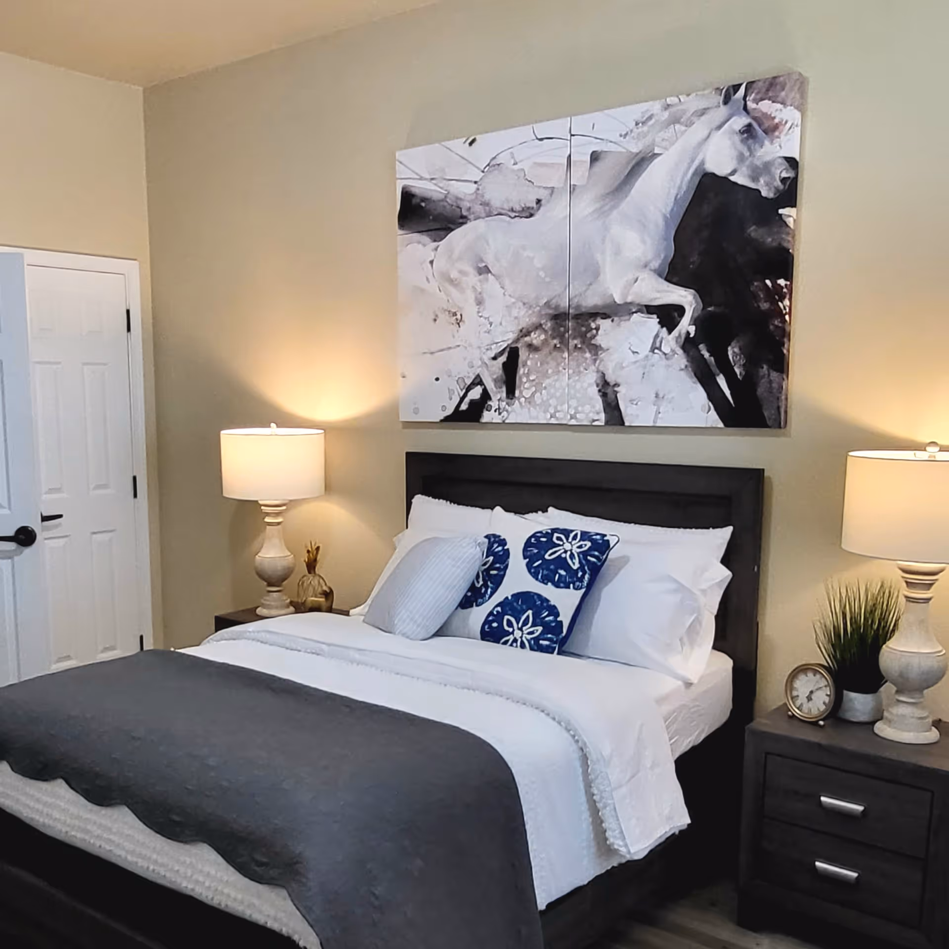 A neatly made bed with white and blue pillows and a dark gray blanket in a bedroom. On either side of the bed are matching nightstands with lamps, a small clock, and a plant. Above the bed is a large artwork of a white horse on a dark and light abstract background. The walls are beige, and there is a white door to the left.