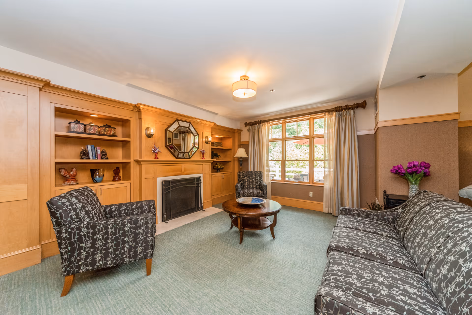 A cozy living room with patterned armchairs and a matching sofa arranged around a wooden coffee table. The room features built-in wooden shelves and cabinetry, a fireplace with a decorative mirror above it, and a large window with curtains letting in natural light.