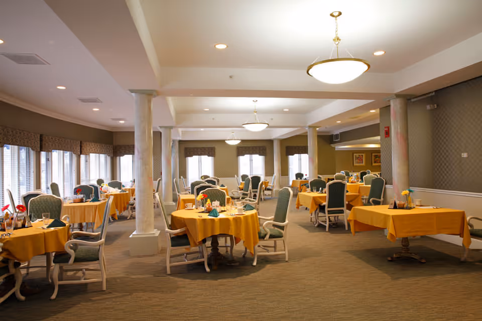 A spacious dining room with multiple round and rectangular tables covered with yellow tablecloths. Each table is set with glassware, napkins, and small flower arrangements. The room has green upholstered chairs, large windows with valances, and several ceiling lights providing warm illumination.