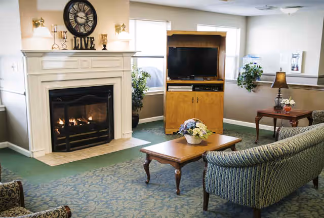 Cozy senior living room with a lit fireplace, TV cabinet, coffee table with flowers, and upholstered seating.