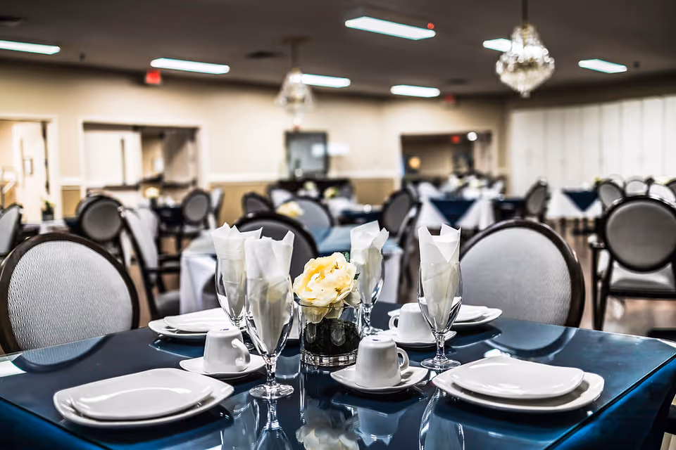 A dining room with tables set for a meal, featuring white plates, cups, folded napkins in glasses, and a small floral centerpiece on a glossy dark table. The room has multiple round tables with cushioned chairs and chandeliers hanging from the ceiling.